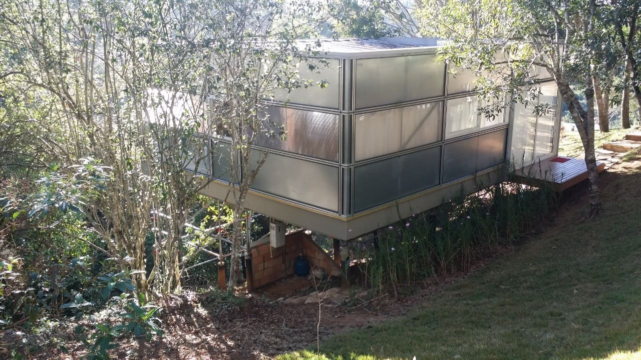 Prédio bege de dois andares sobre palafitas em uma encosta arborizada, com telhado inclinado e árvores ao redor.