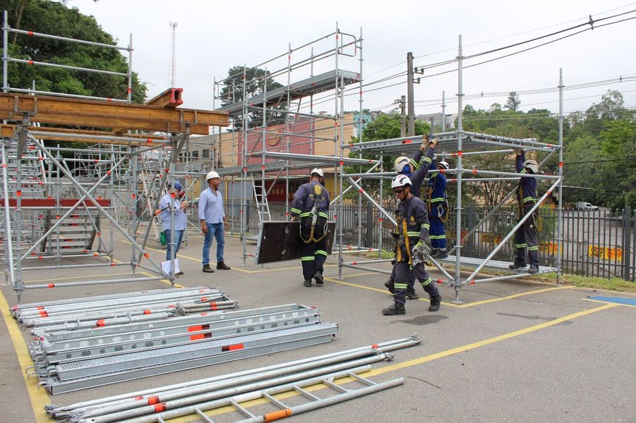 Operários com equipamentos de segurança estão próximos a andaimes e tubos de metal em um canteiro de obras.
