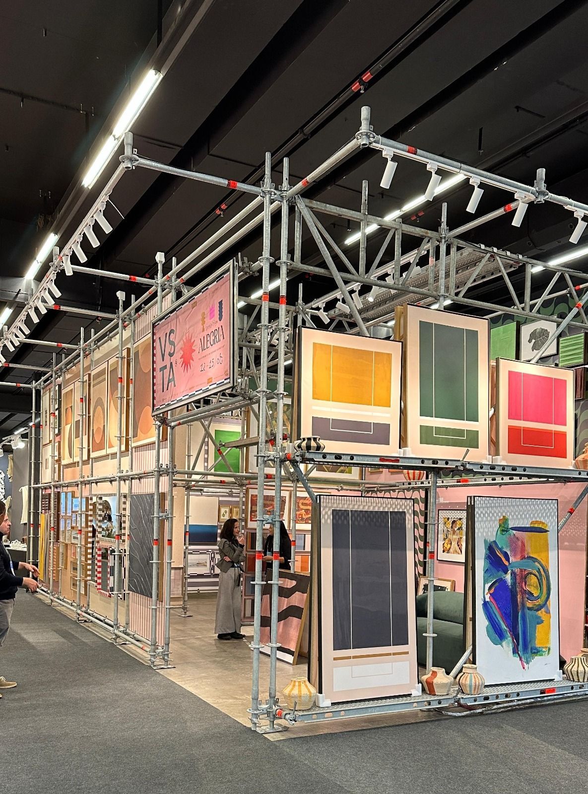 Exposição de arte em ambiente fechado com gravuras coloridas emolduradas em prateleiras de metal sob luzes brilhantes.