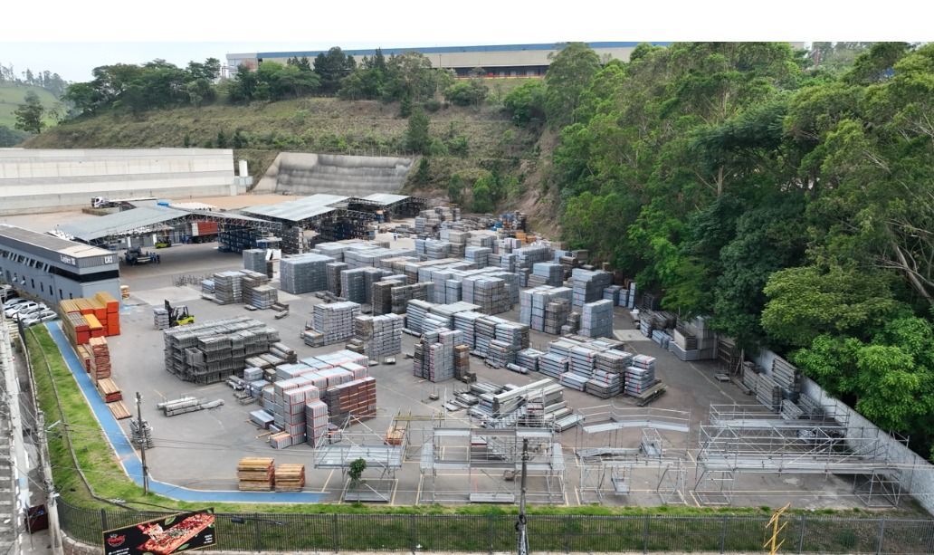 Grande pátio industrial ao ar livre com pilhas de blocos de concreto e equipamentos ao lado de árvores.
