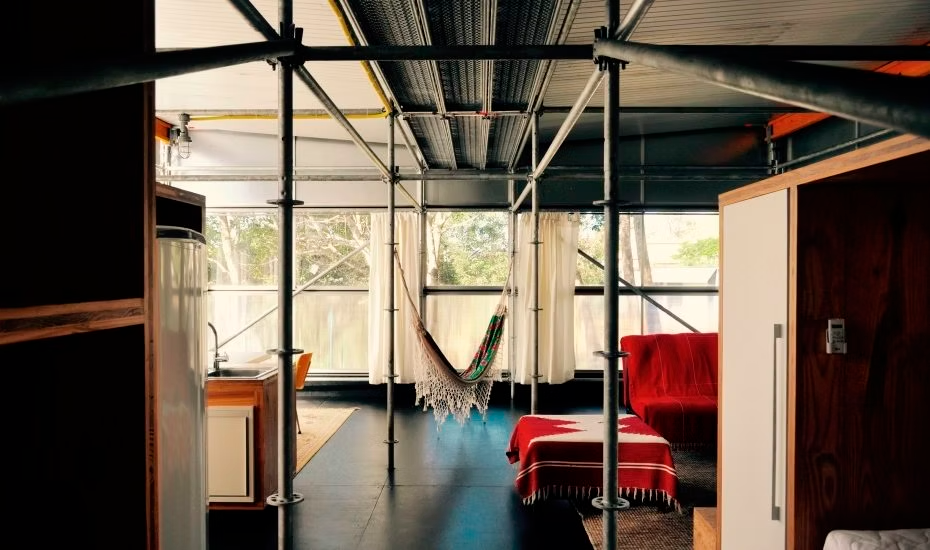 Quarto iluminado com estrutura metálica, cadeira de balanço suspensa e sofá vermelho junto a grandes janelas.