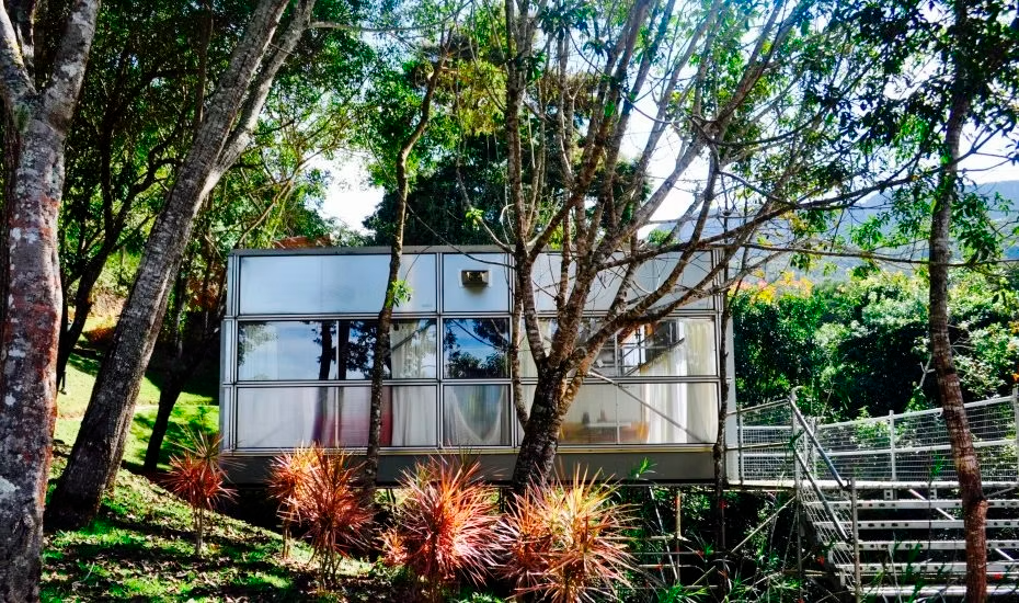 Pequeno prédio branco com grandes janelas em um jardim arborizado, com escadas e plantas vermelhas na frente.