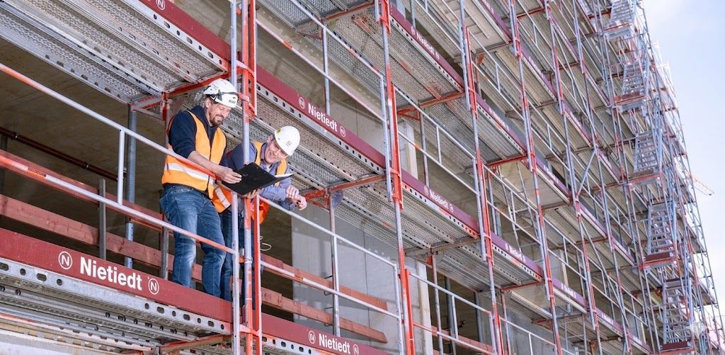 Operários com coletes de segurança laranja em andaimes ao lado de um prédio em construção.