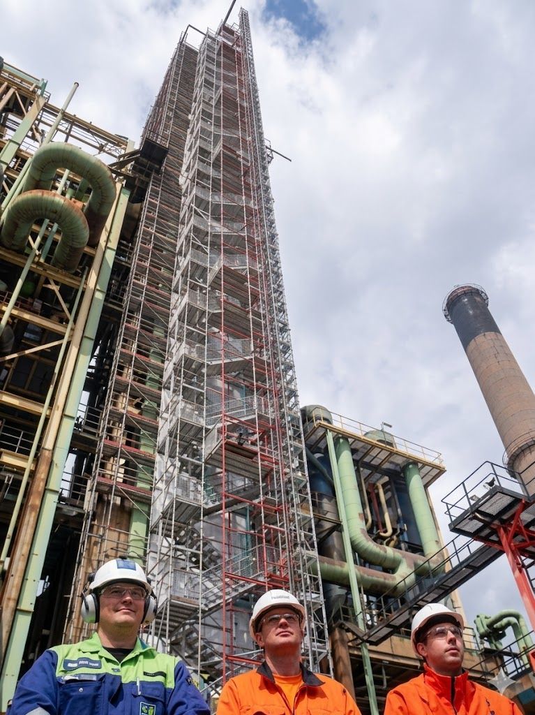Três operários com capacetes de segurança estão em frente a uma imponente estrutura industrial em construção.