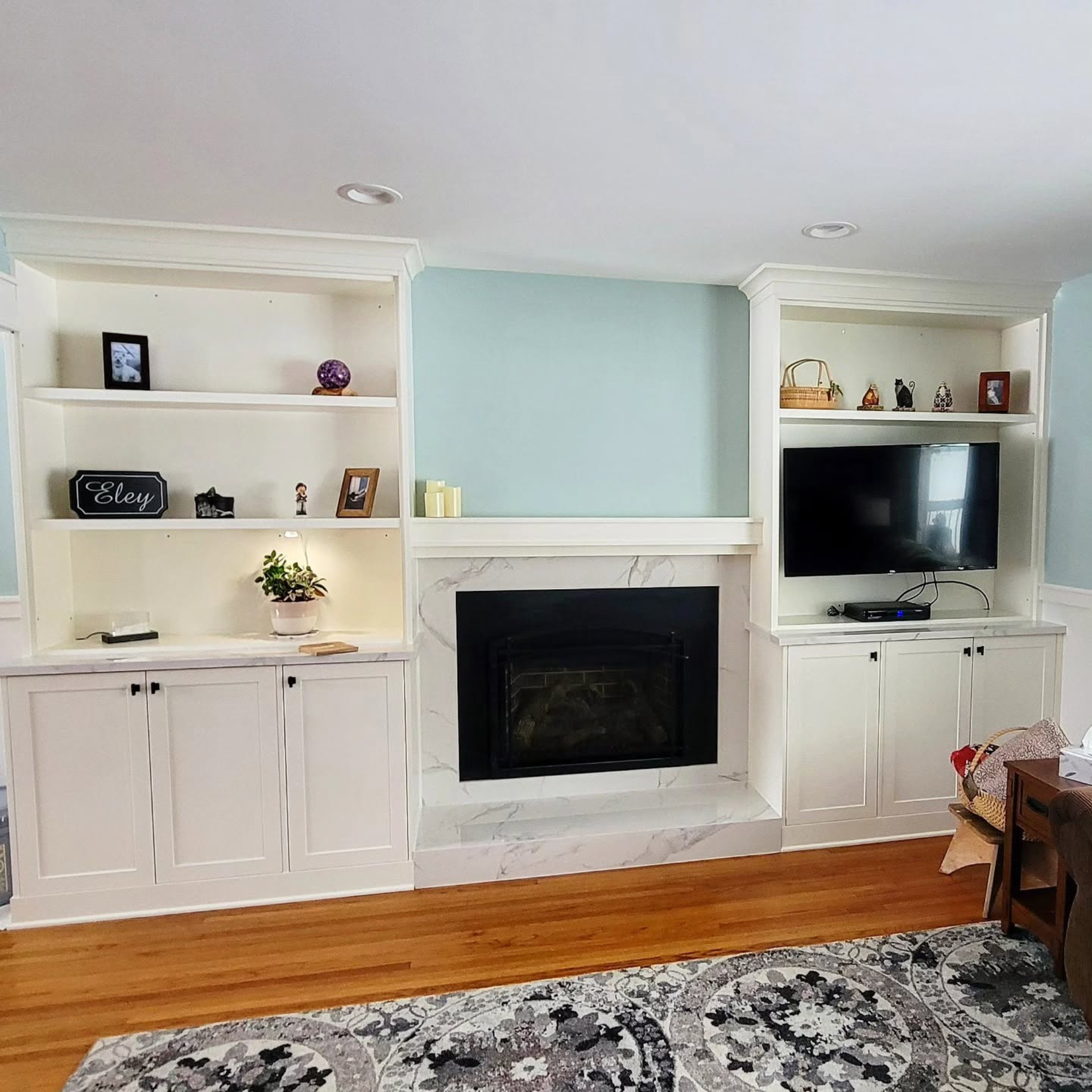 Living room built-ins with white cabinets, a fireplace, TV nook, and blue accent wall above wood floors and rug