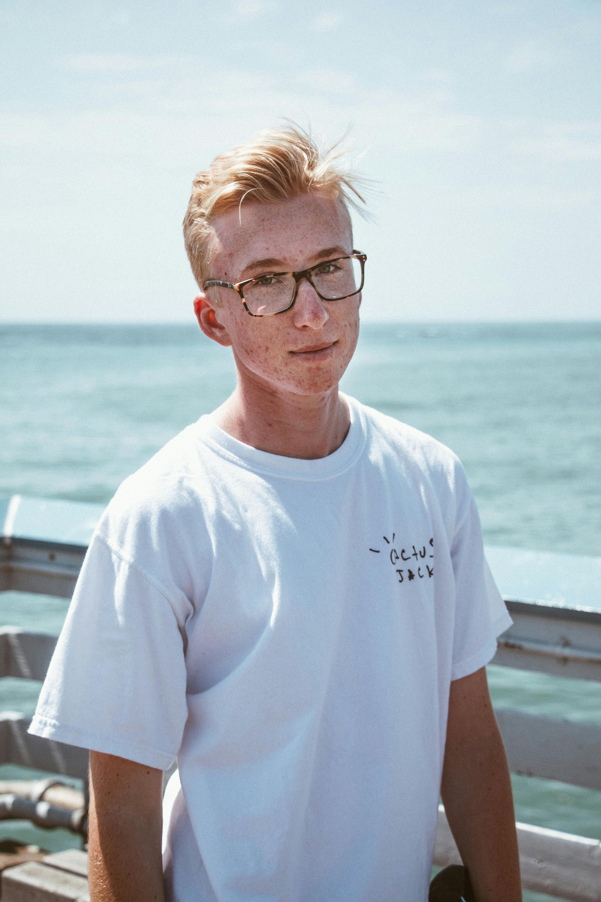 Een jongeman met een bril en een wit overhemd staat op een pier met uitzicht op de oceaan