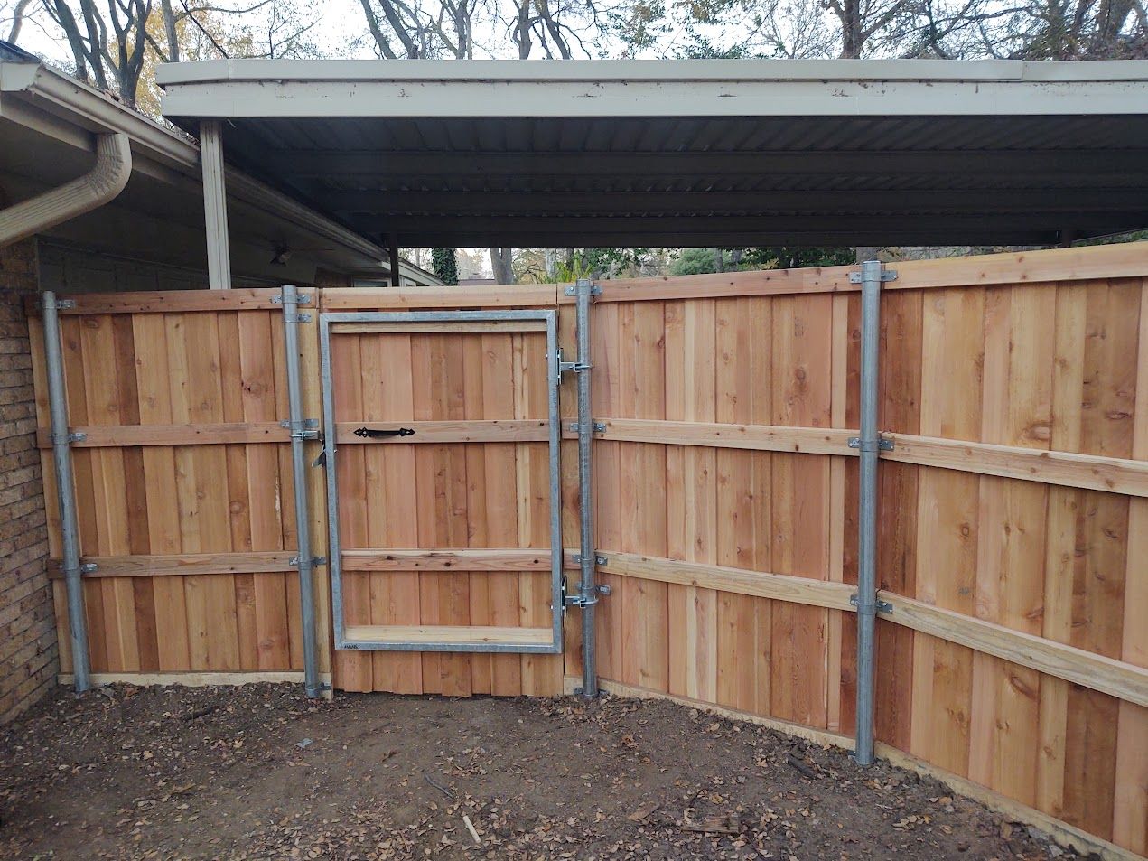 Wooden fenced yard with a metal gate under a corrugated roof, viewed from inside.