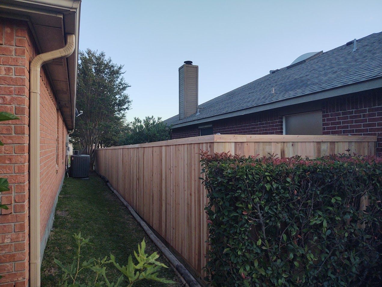Narrow side yard between brick house and fence, with hedge and blue-tiled roof at dusk