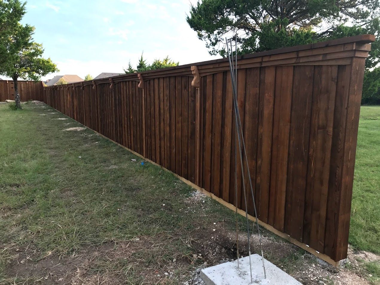 Long brown wooden fence along a grassy yard beside a sidewalk, with trees in the background.
