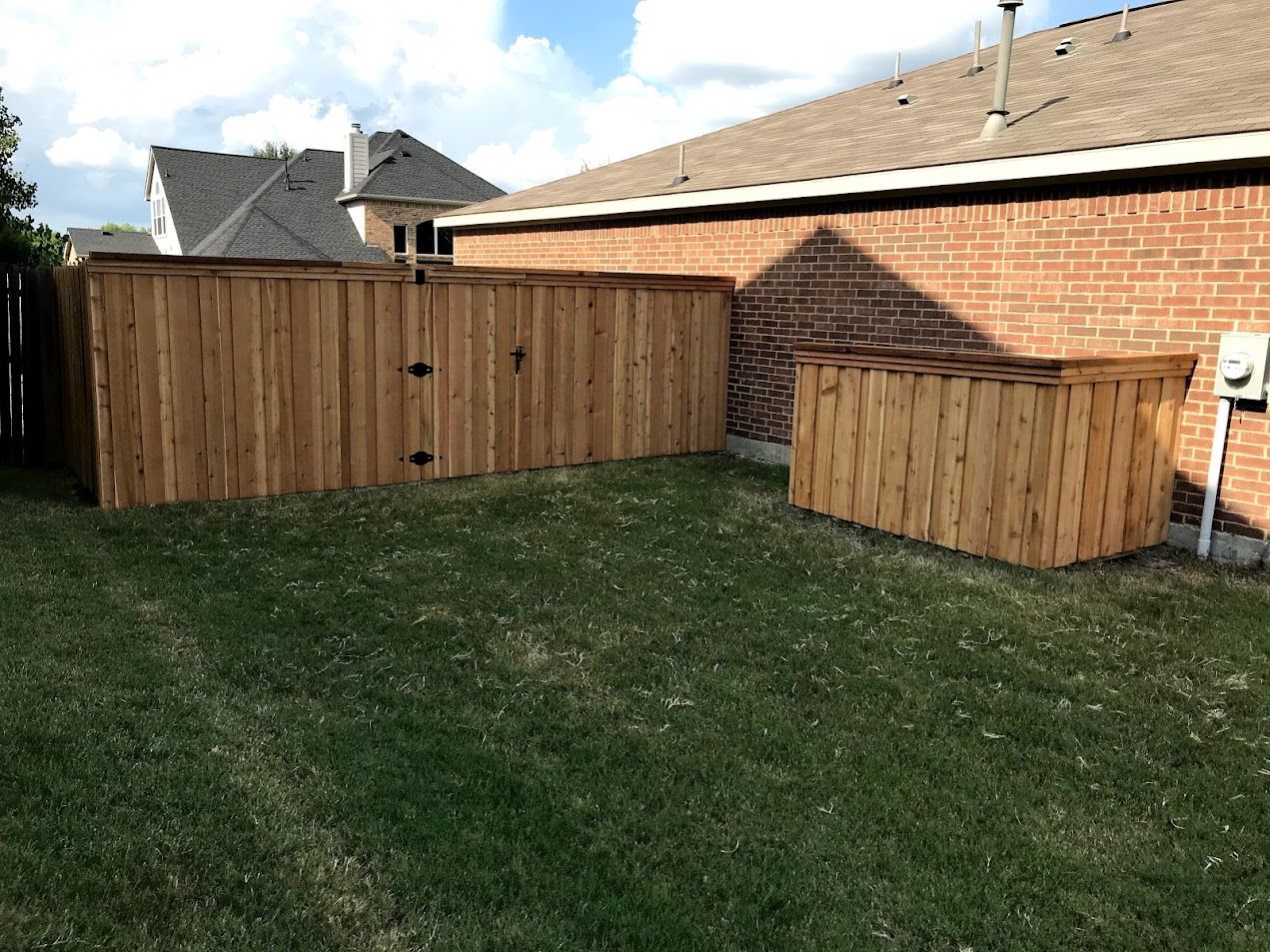 Backyard with two wooden fences, brick house wall, and grass lawn under a partly cloudy sky
