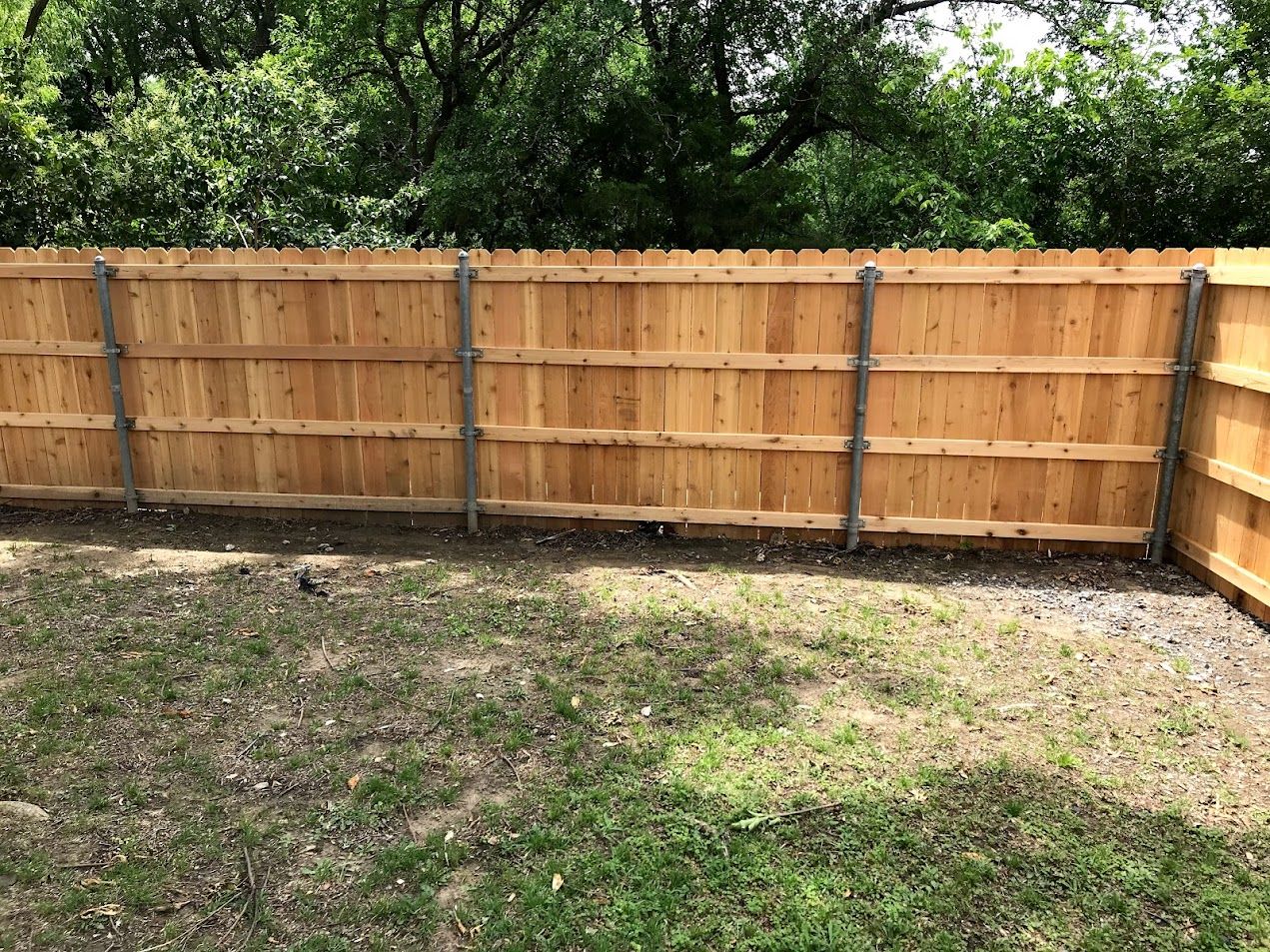 Wooden backyard fence bordering a patchy grass yard with trees behind it