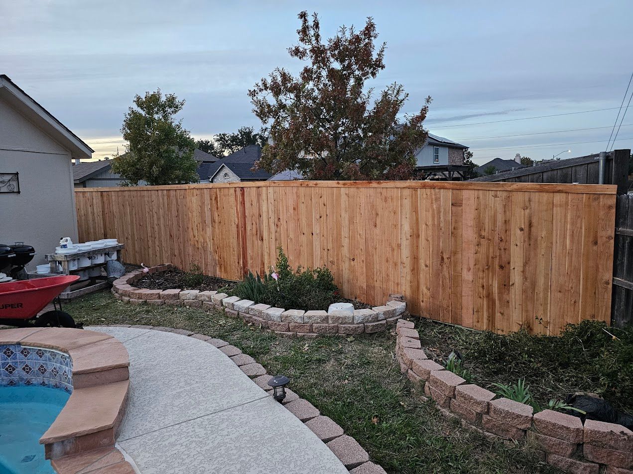 Backyard garden with a wooden fence, curved stone edging, and a concrete patio at dusk