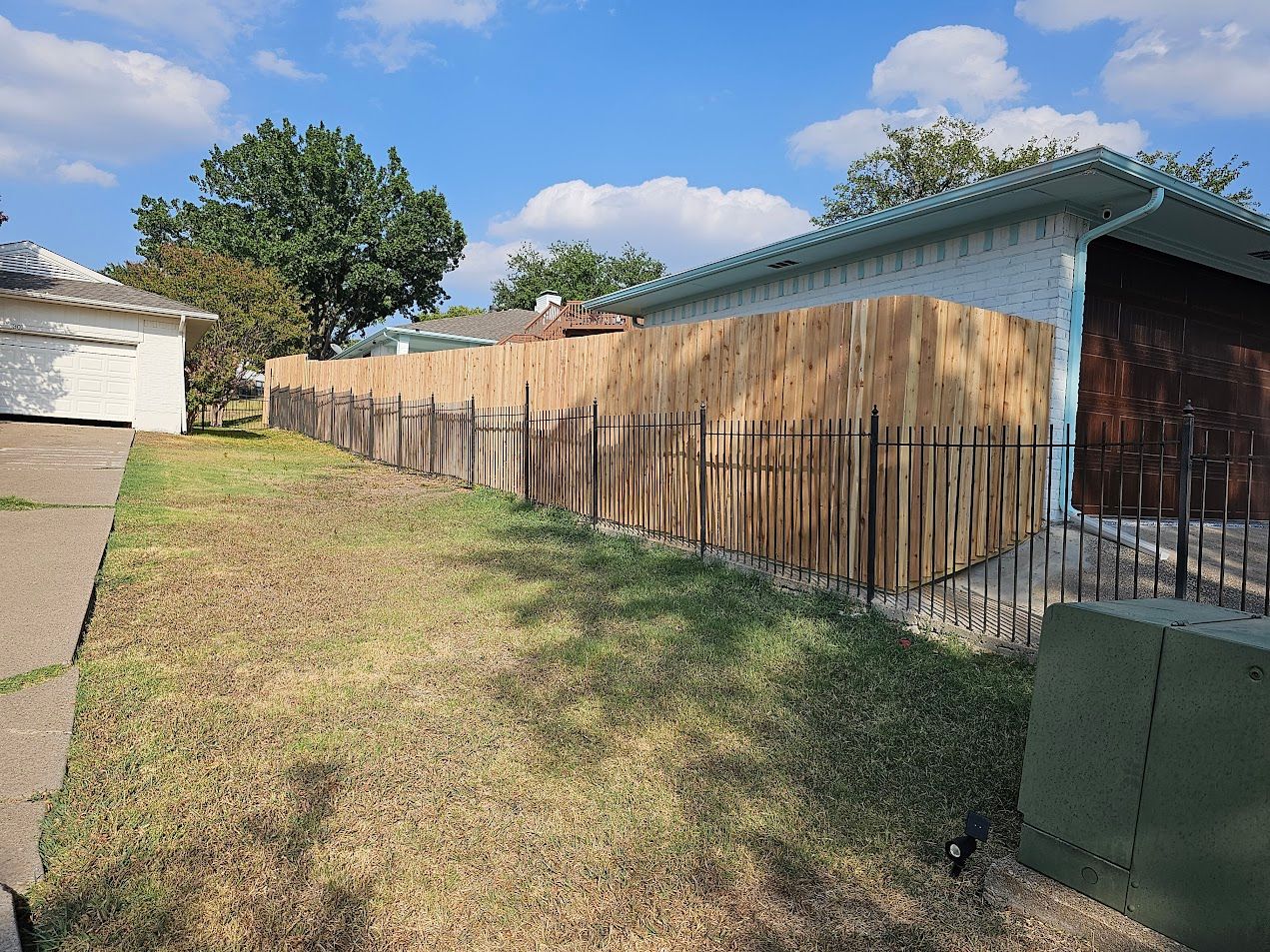 Backyard with a wooden fence, grassy yard, and a tree beside a house under a blue sky.