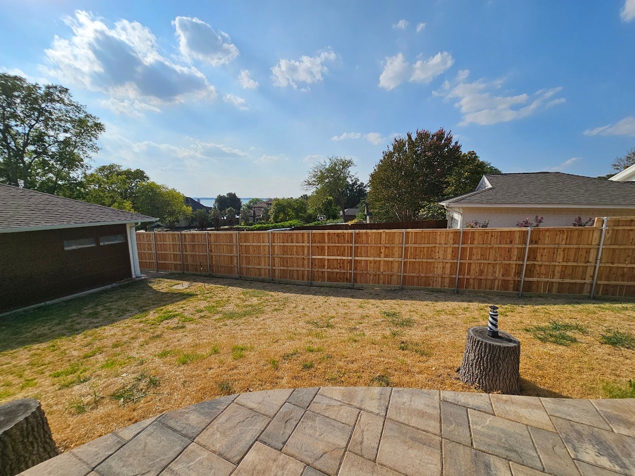 Backyard with wooden fence, patio pavers, dry grass, and a small fountain under a blue sky