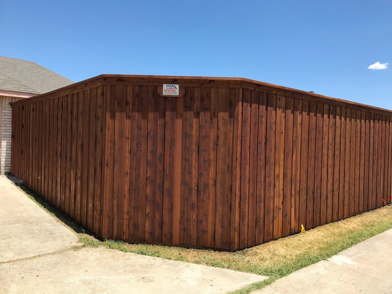 Dark wooden fence surrounding a corner of a yard beside a paved path under a blue sky