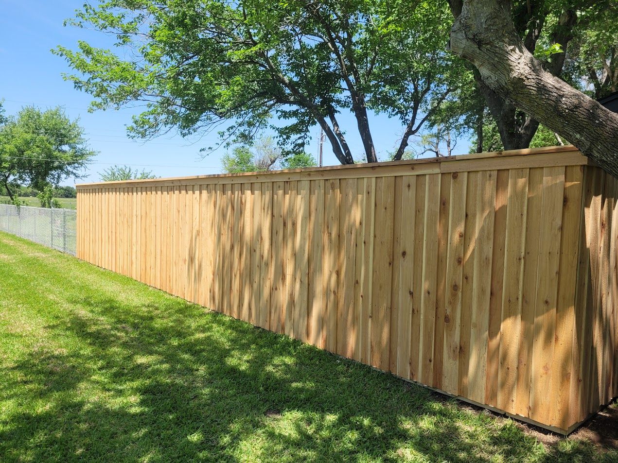 Wooden privacy fence along a grassy slope with trees and blue sky overhead