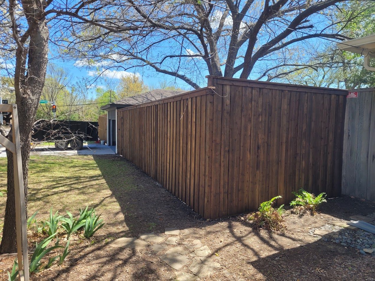Wooden fence and shed in a sunny yard with trees and a parked vehicle in the background