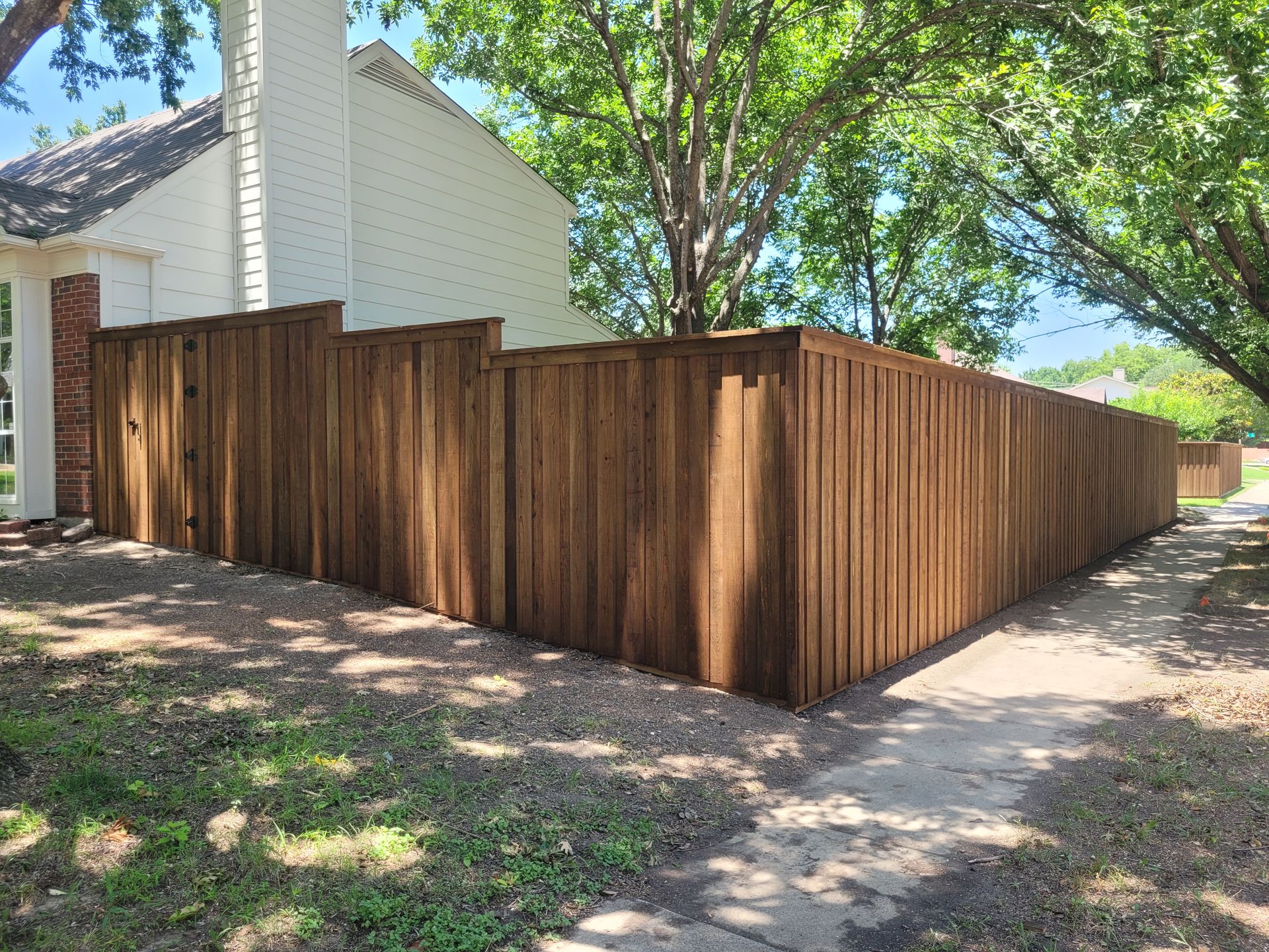 Wooden privacy fence around a house corner beside a sidewalk and trees in daylight