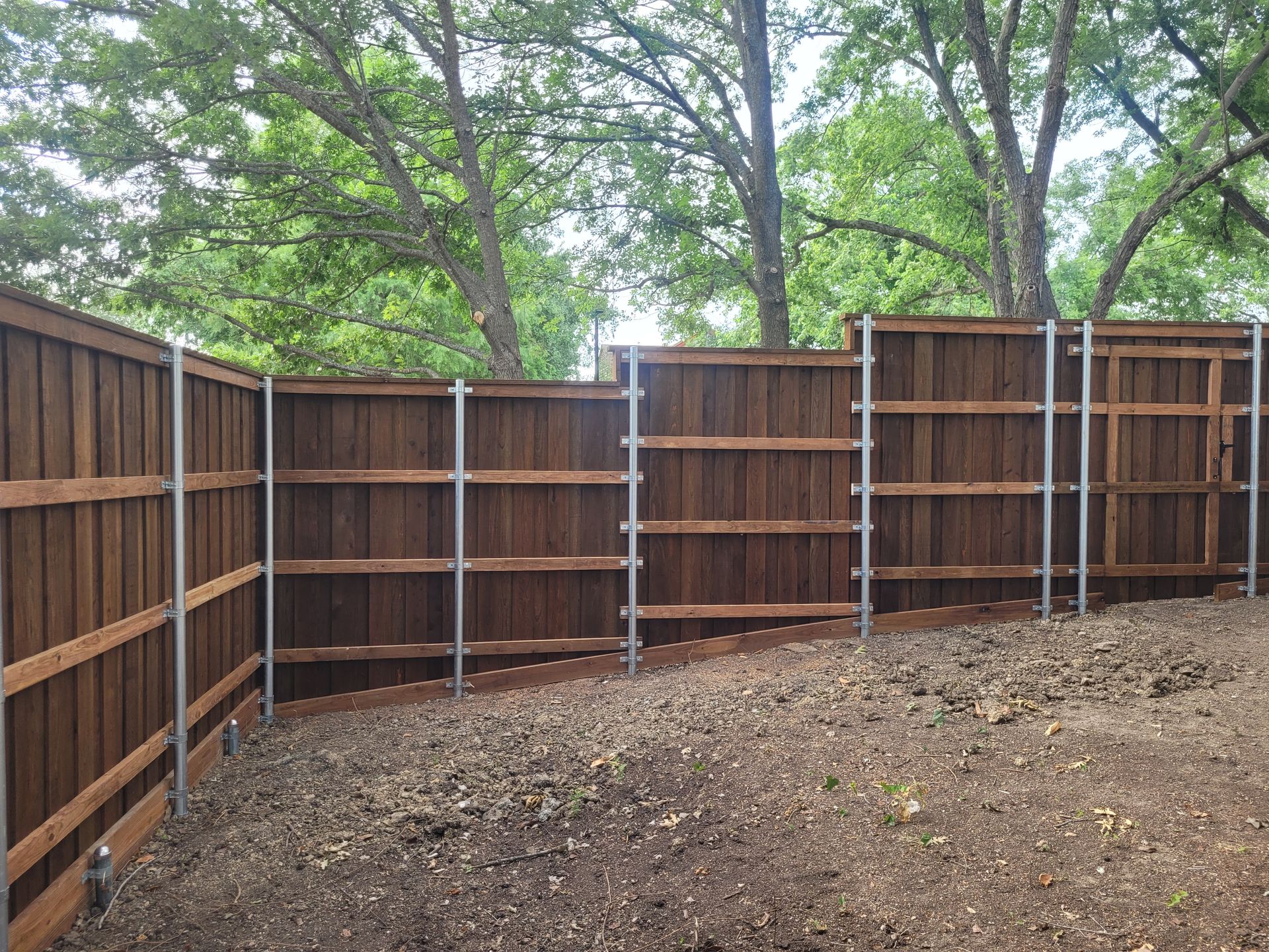 Brown wooden fence around a bare dirt yard with trees in the background