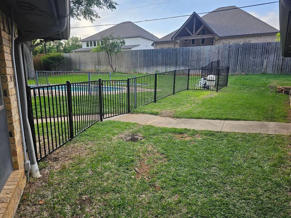 Backyard with green lawn, black fence, concrete path, and patio chair beside a wooden privacy fence.