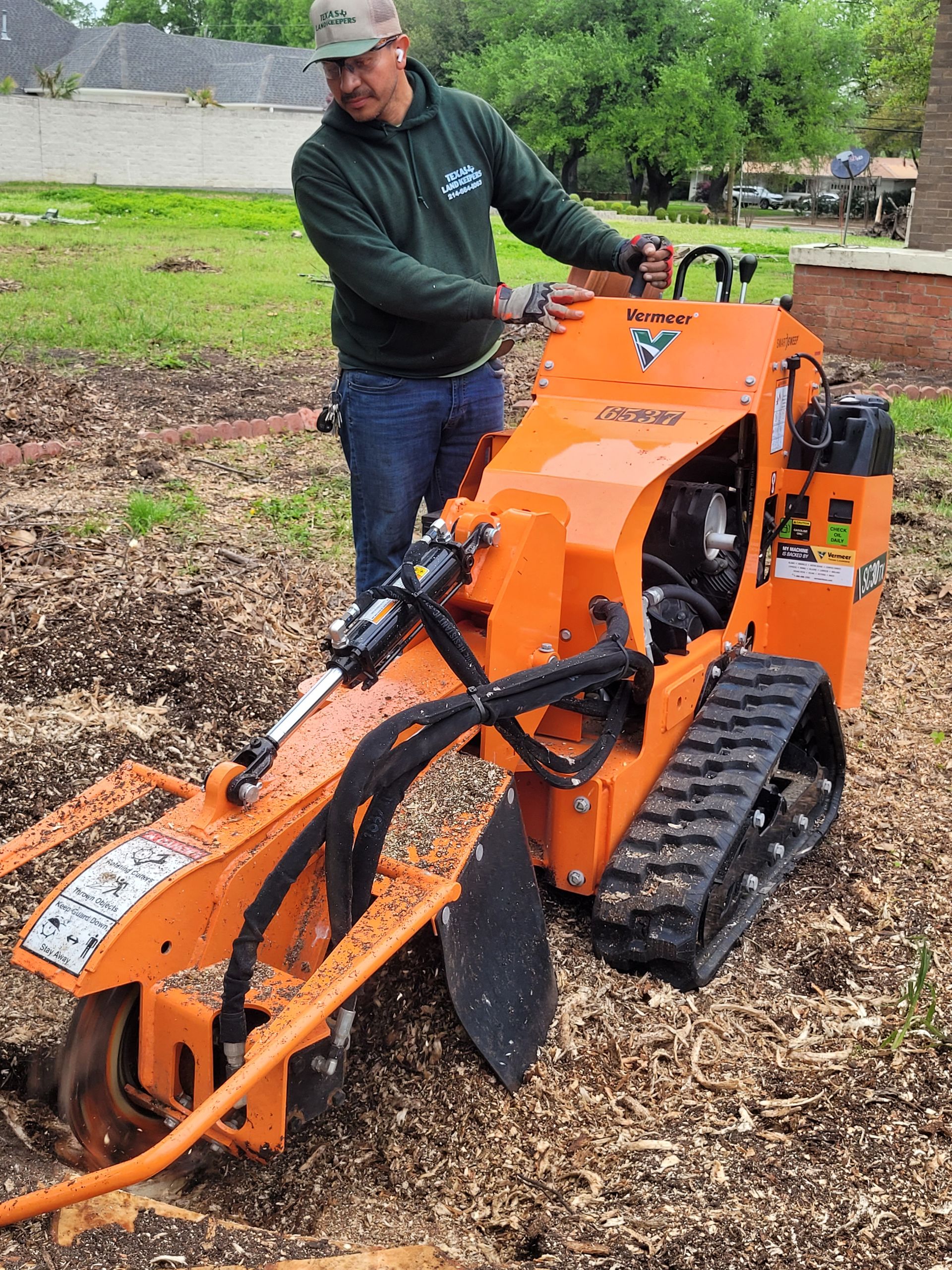 Person operating an orange tracked trenching machine on a dirt and woodchip path outdoors