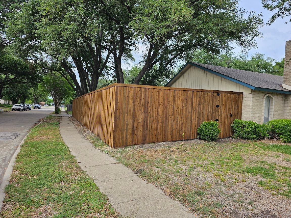 Residential street corner with a wooden privacy fence, sidewalk, trees, and a house behind it.