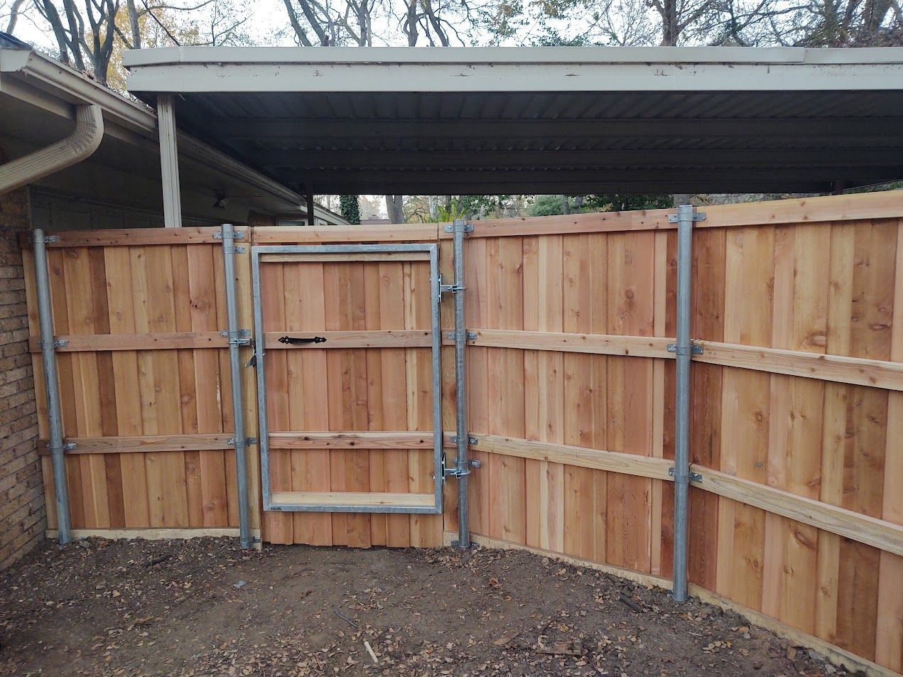 Wooden fenced enclosure with a metal gate and gravel ground under a corrugated roof