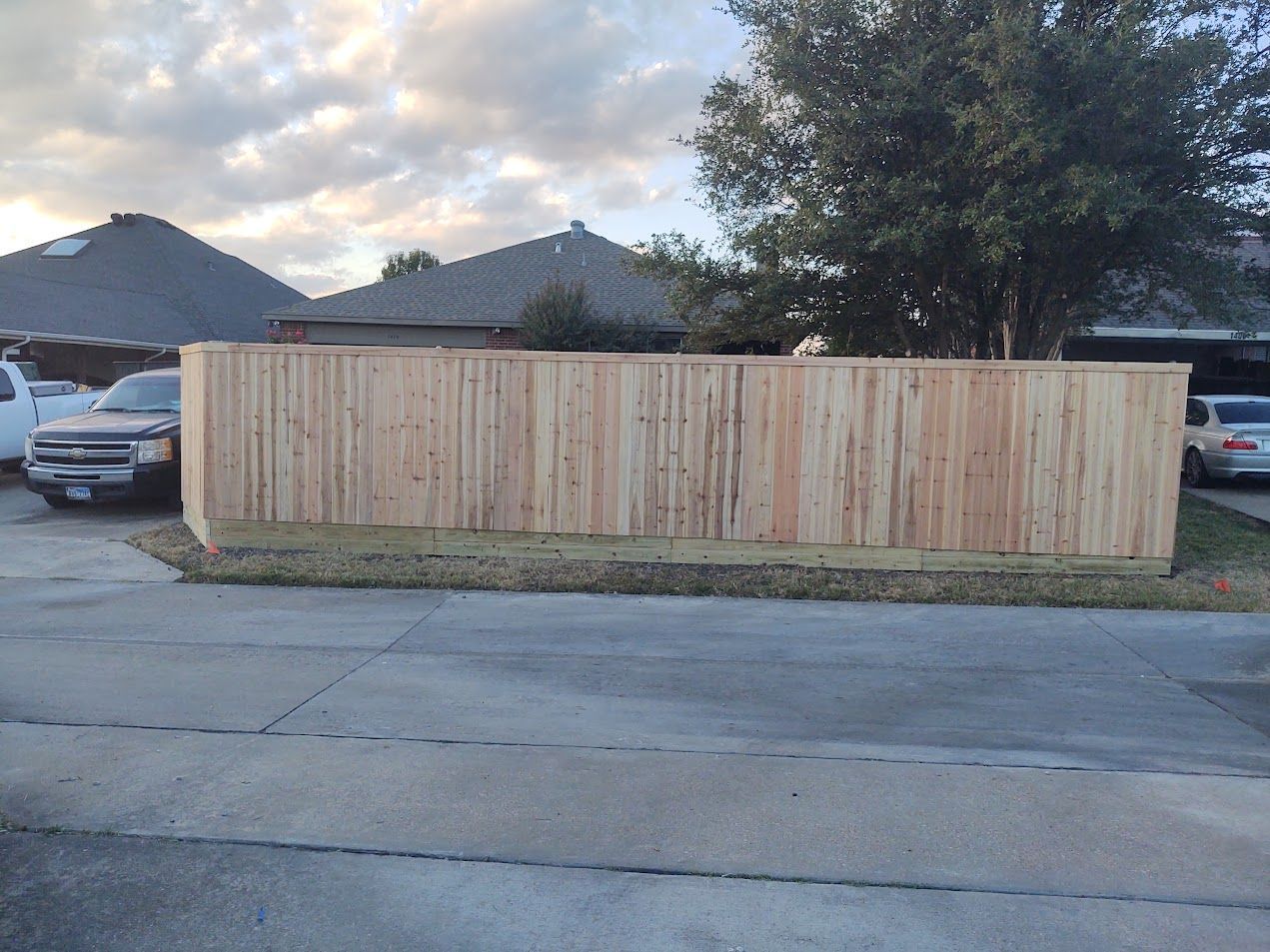 Wooden privacy fence along a residential street at dusk, with parked cars and houses in the background.