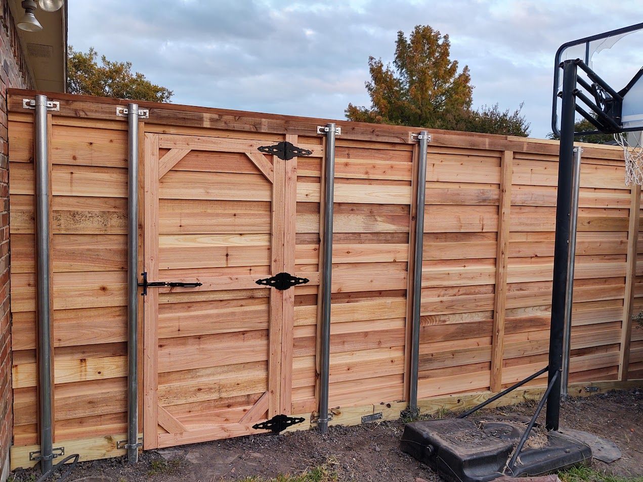 Wooden backyard fence with a central gate beside a basketball hoop pole under cloudy sky