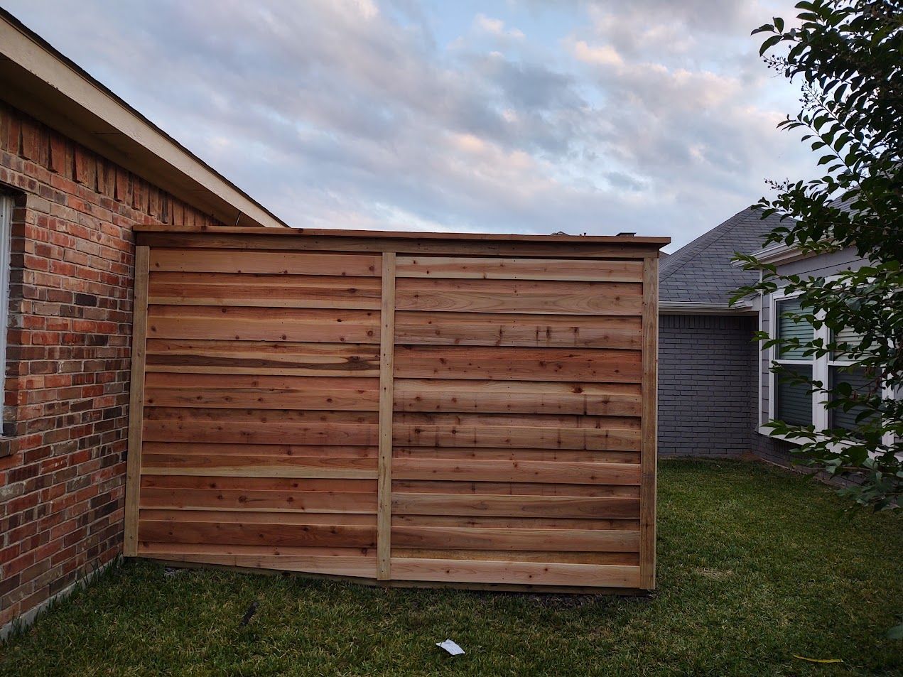 Wooden privacy fence panels beside a brick house in a grassy backyard at dusk