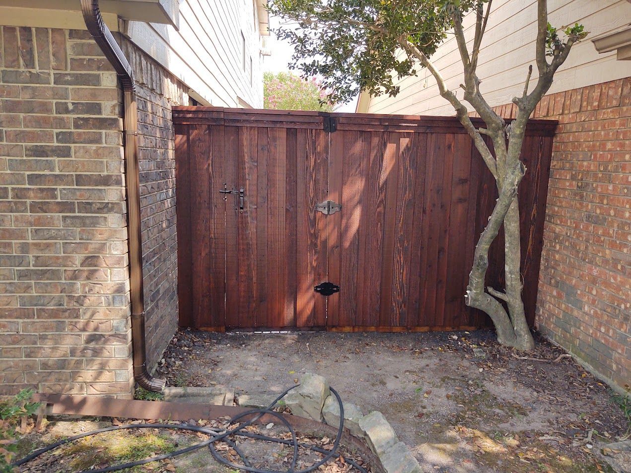 Wooden double gate between brick walls, with a tree beside it and a paved driveway in front.