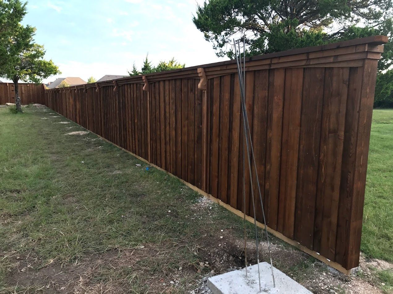 Long brown wooden fence along a grassy slope in a yard, with trees and a sidewalk nearby