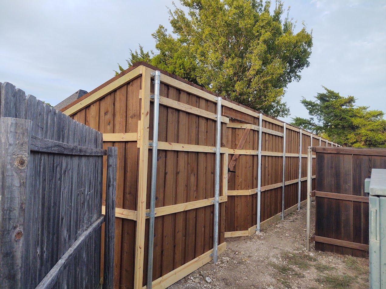 Wooden fence under construction along a backyard path, with unfinished posts and rails beside a weathered fence