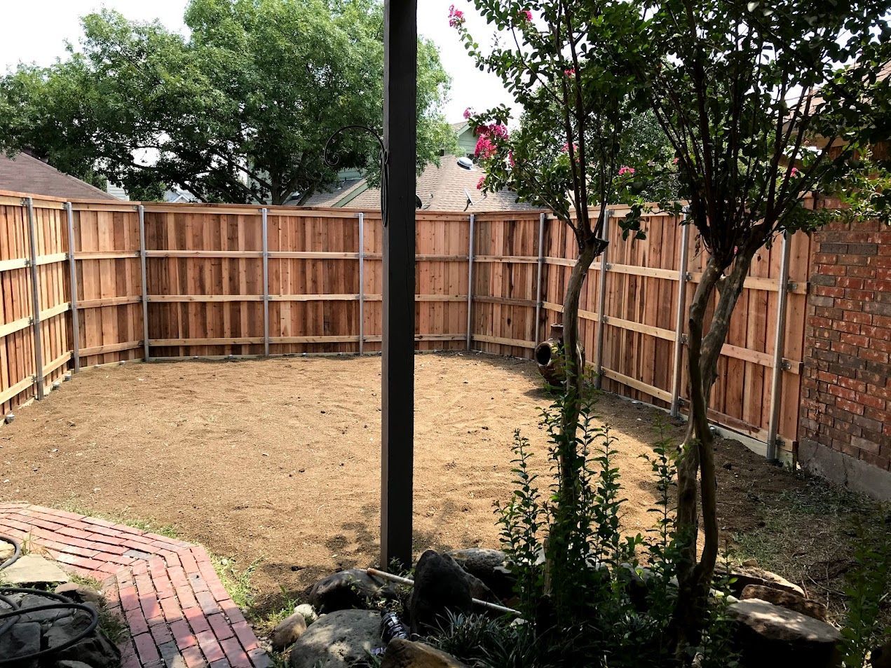 Wooden-fenced backyard with a dirt yard, trees, and a small rock garden in the foreground