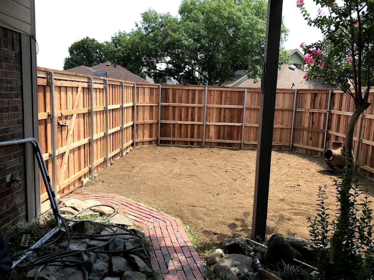 Backyard with a new wooden fence, bare ground, and brick edging under trees