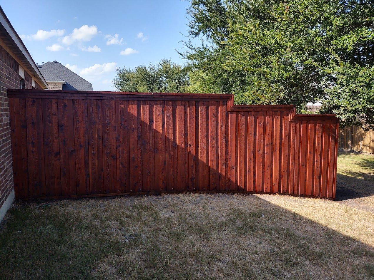 Red wooden fence and gate in a sunny backyard beside a brick house and tree.