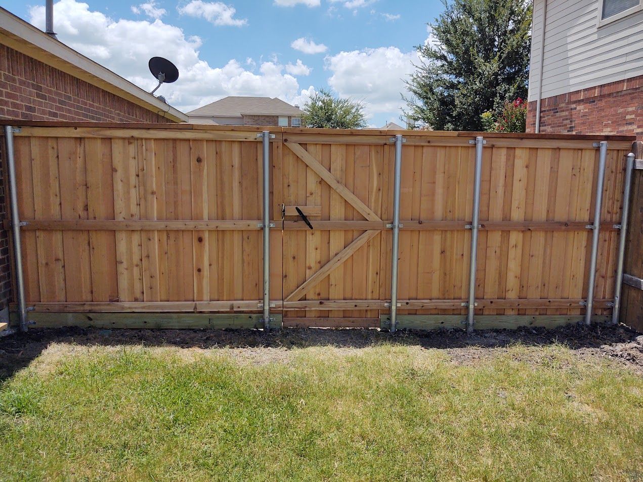 Wooden backyard fence with double gate, set between two brick houses under a partly cloudy sky