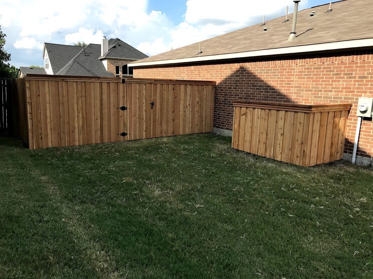 Backyard with grass, wooden fence, and brick house under a partly cloudy sky