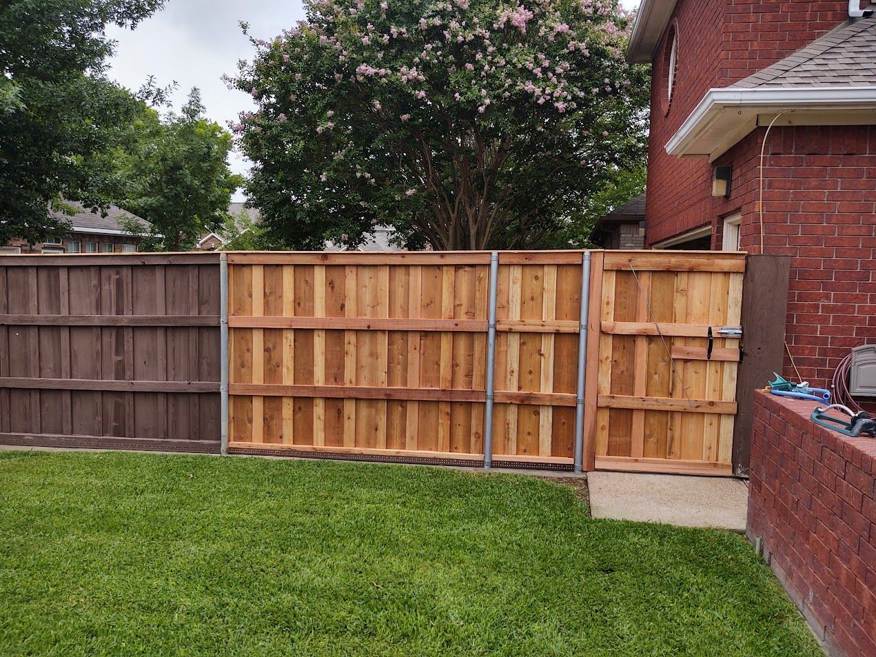 Backyard with green lawn, wooden fences, a brick house wall, and a tree in the background