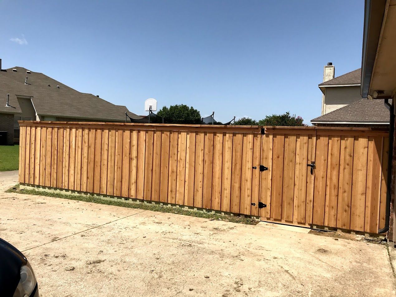 Wooden fence and gate along a gravel driveway beside suburban houses under a clear sky