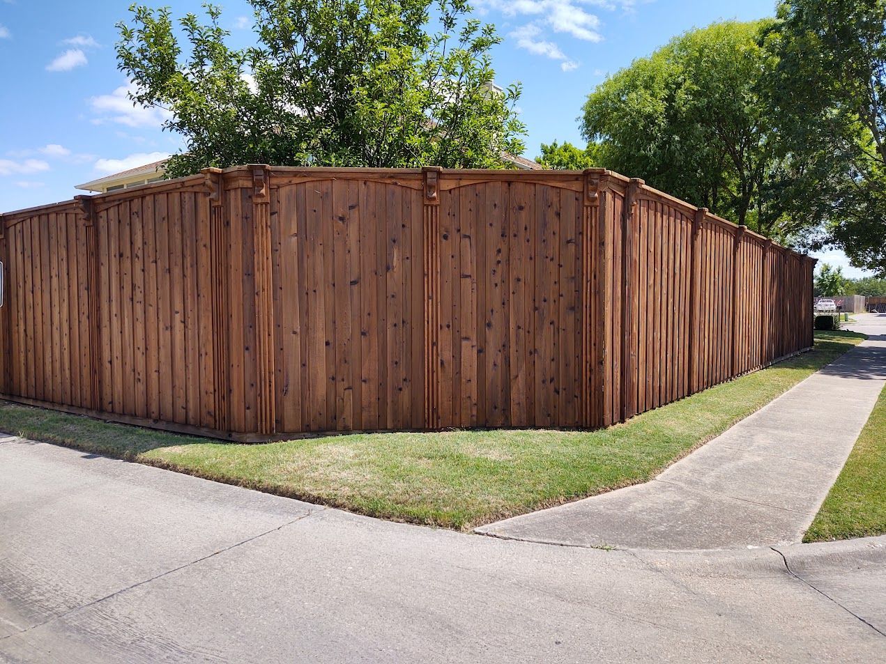Wooden privacy fence along a street corner beside a sidewalk and grass, with trees in the background