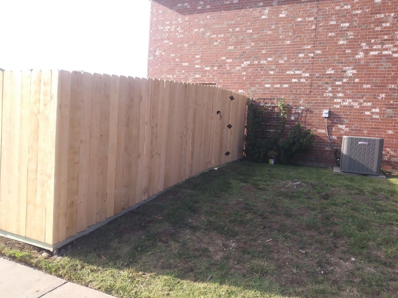 Wooden privacy fence along a grassy yard beside a brick wall, with an AC unit in the corner.