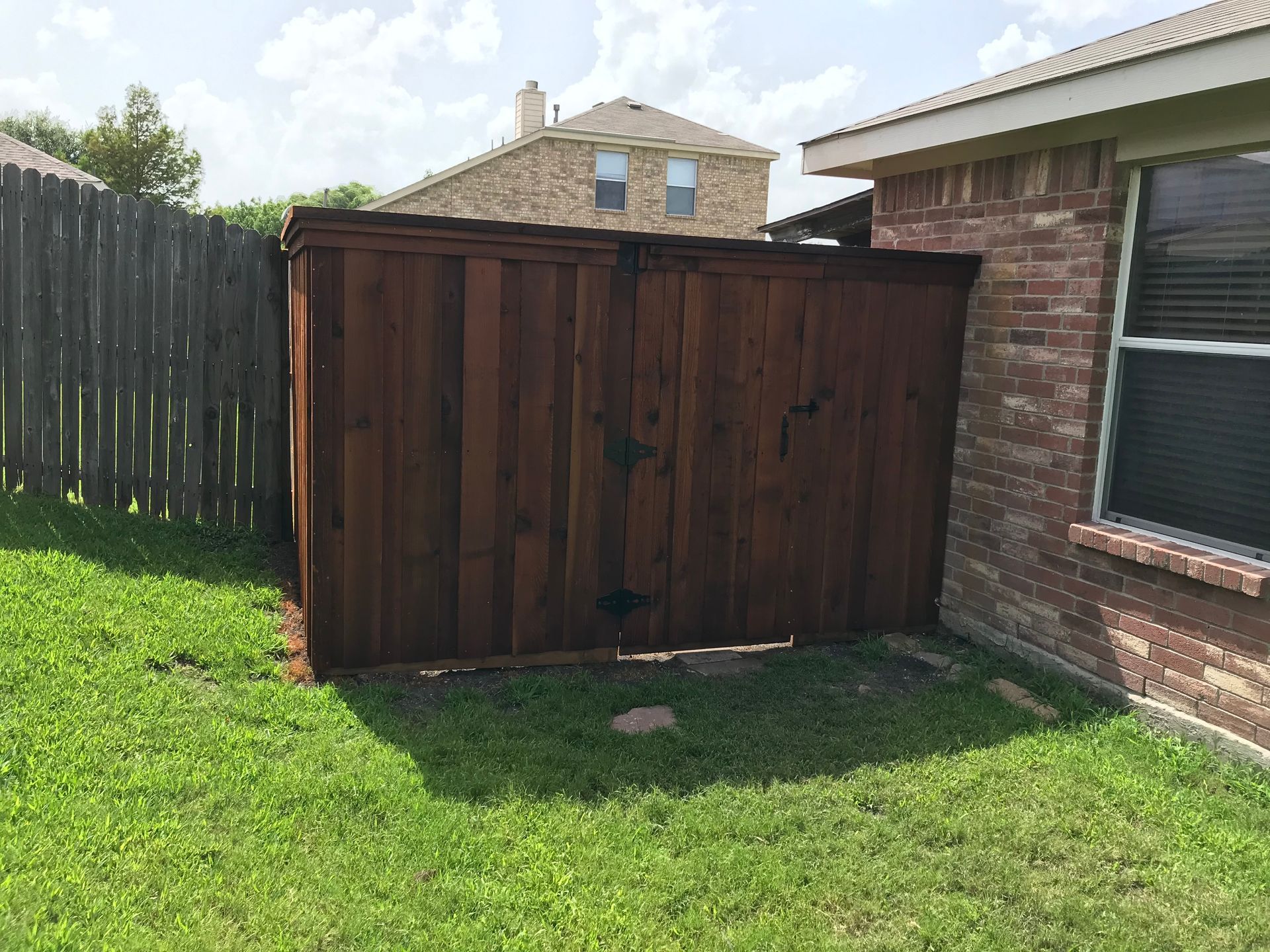 Large dark wooden shed beside a brick house in a grassy backyard with a black fence.