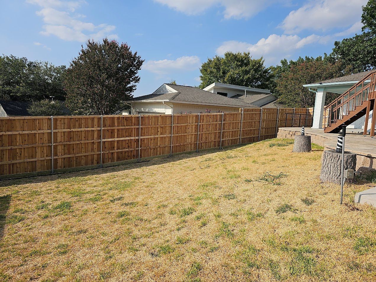 Backyard with dry grass, wooden fence, trees, and a small stone patio under a blue sky