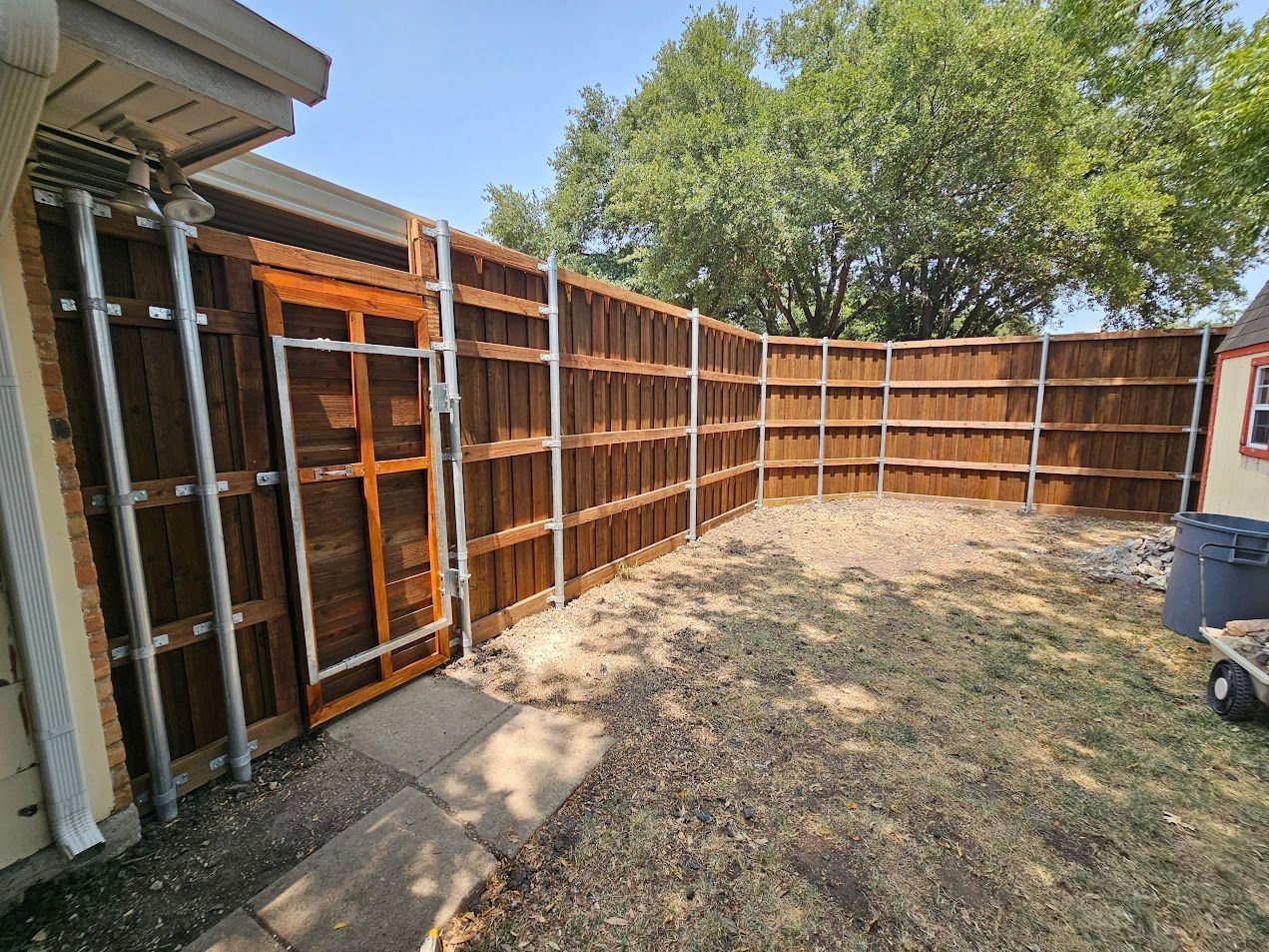 Backyard with a long wooden fence, dry grass, and a small patio area beside a house