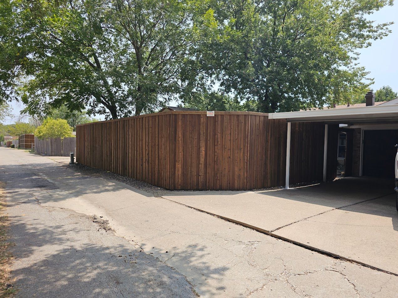 Wooden fence and carport beside a driveway under trees on a sunny day
