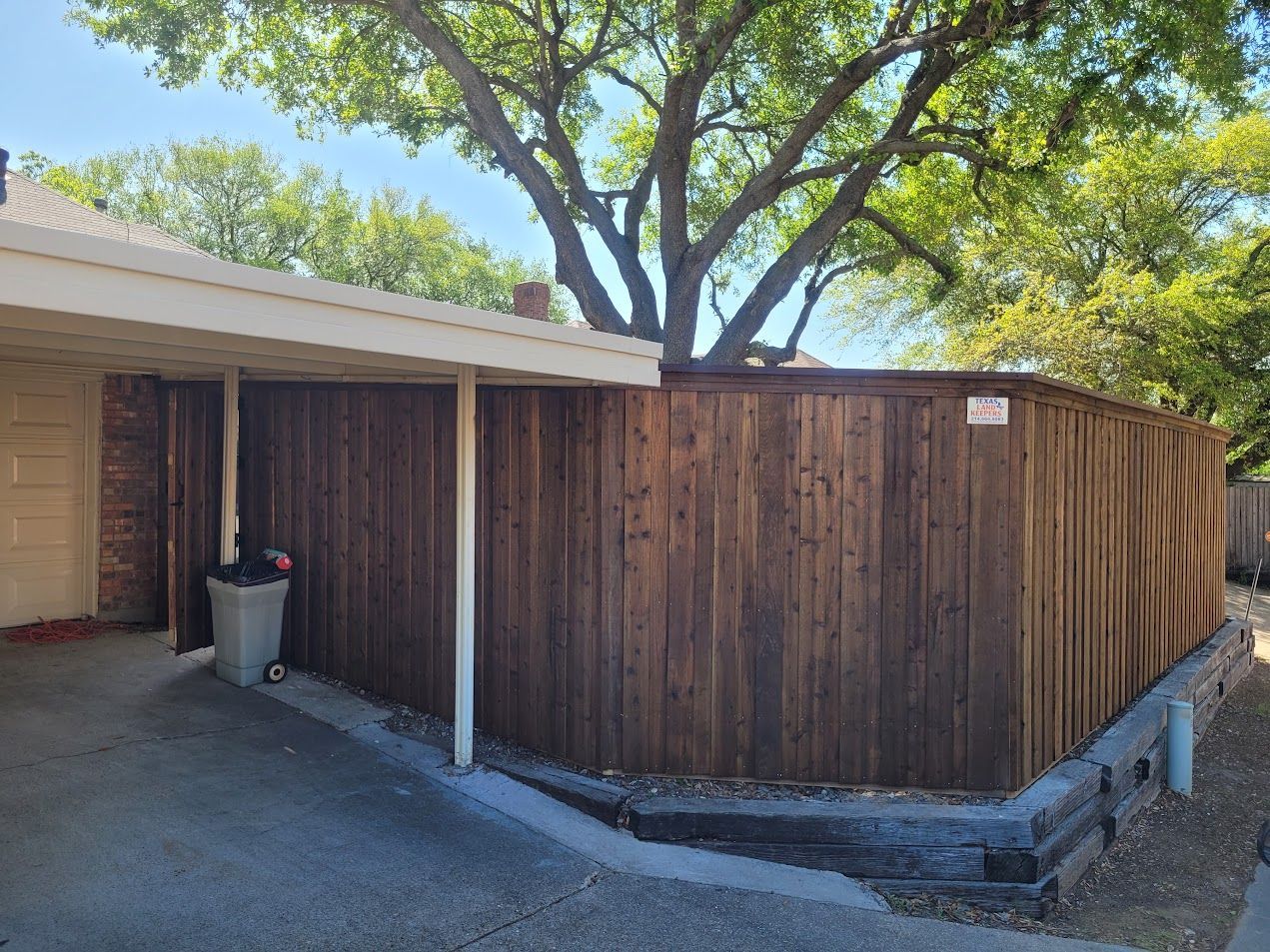 Wooden privacy fence beside a house with a driveway and a trash can under trees