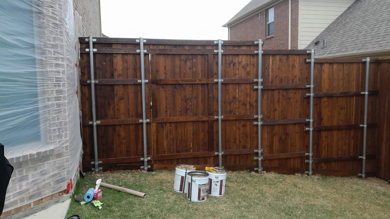 Backyard with wooden fence, grass, and paint cans near a house and neighboring brick home