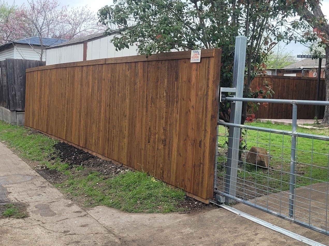 Wooden fence beside a chain-link gate along a sidewalk and dirt strip.