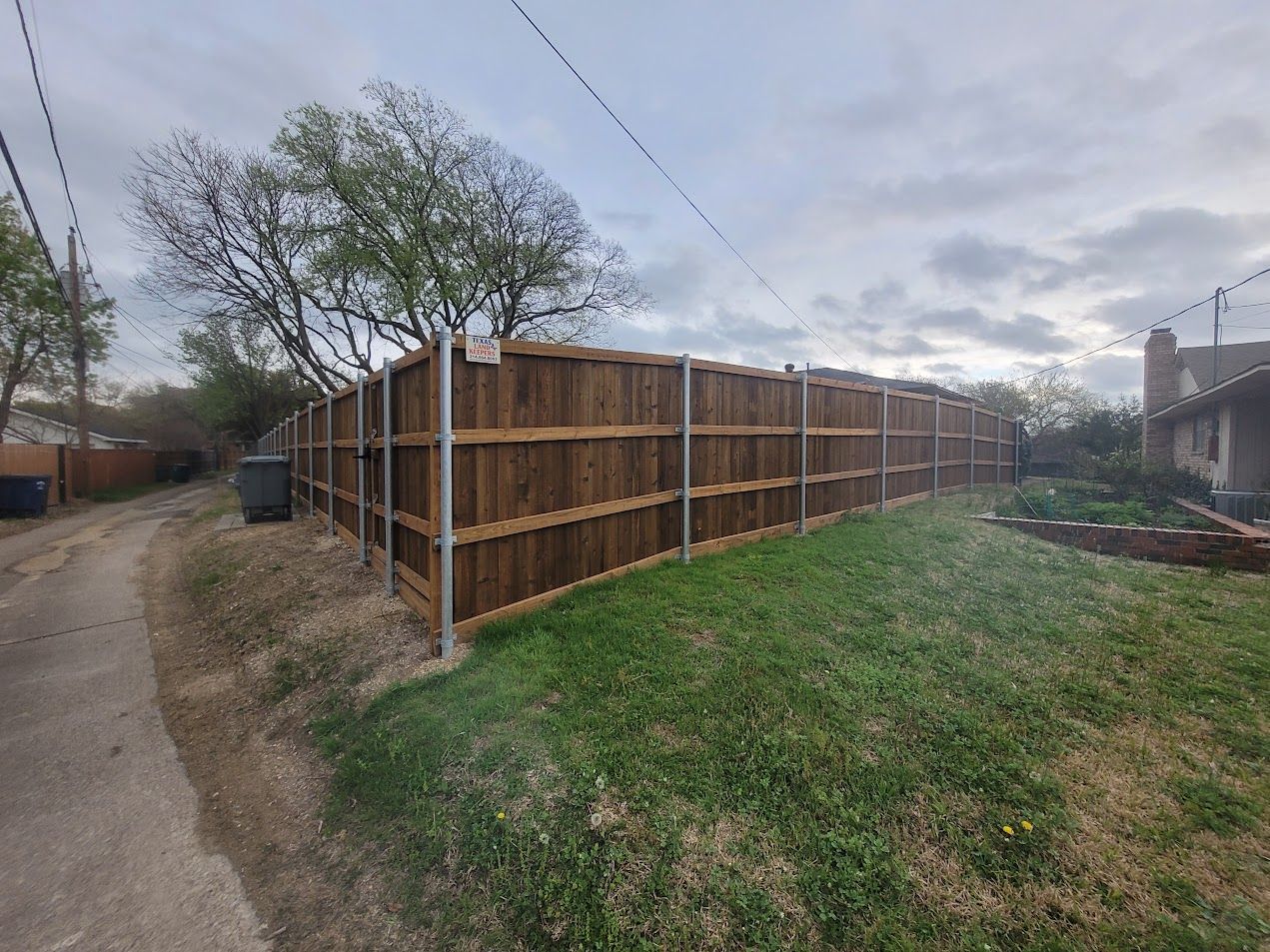 Wooden privacy fence along a grassy yard beside a dirt path under a cloudy sky